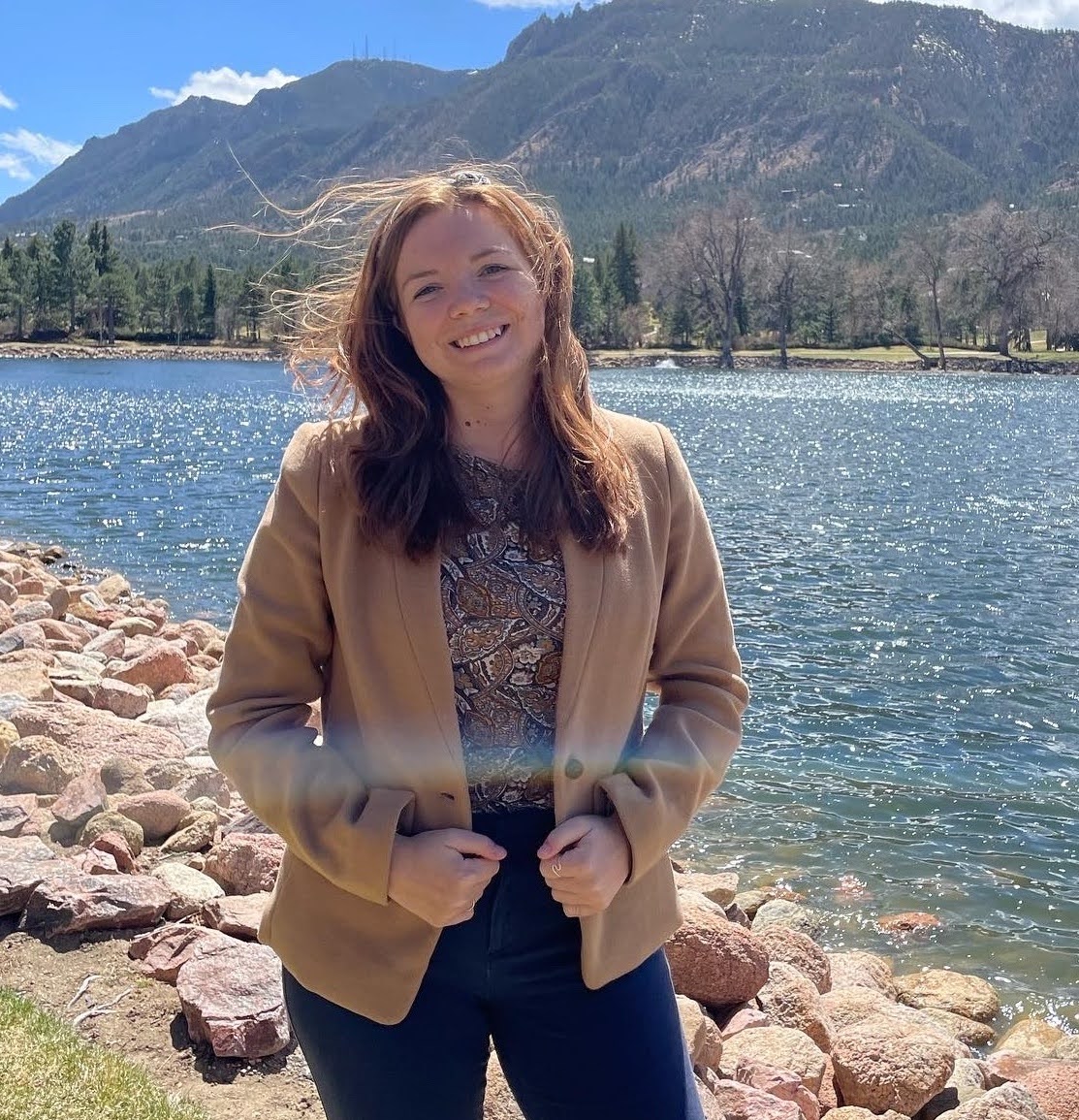 A headshot of Lindsey, standing in front of a lake and wearing professional attire.